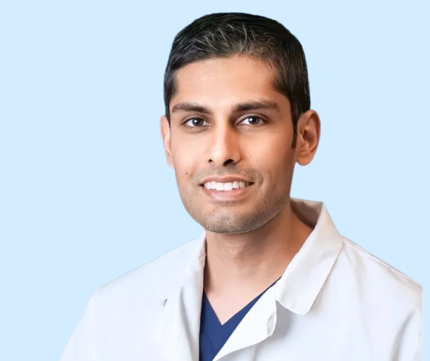 Male dentist in white coat and blue scrubs smiling against light blue background.