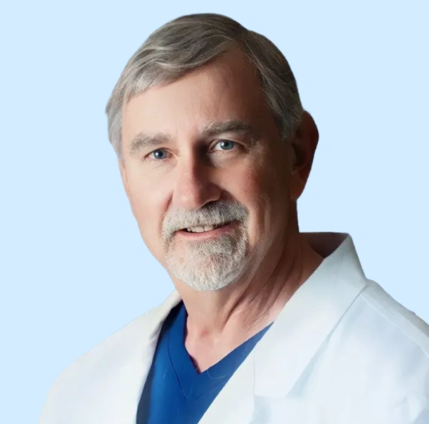 Professional headshot of male dentist wearing white coat and blue scrubs against light blue background.