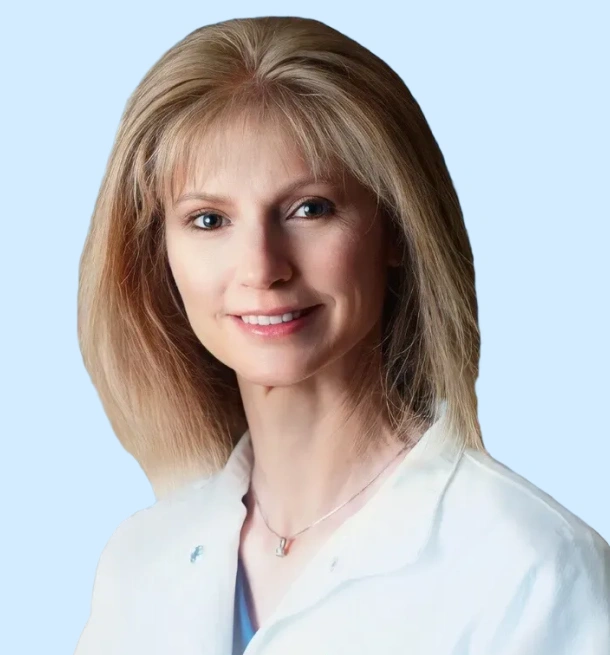 Professional headshot of female dentist in white coat against light blue background.