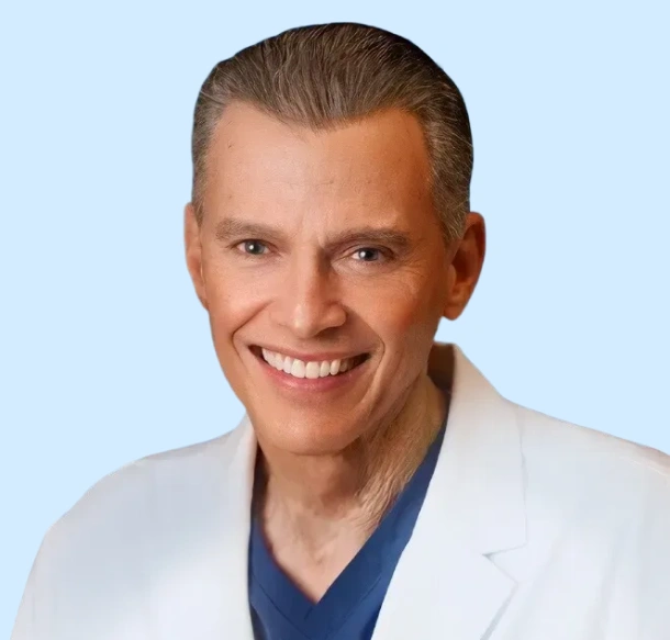 Professional headshot of male dentist in white coat and blue scrubs, smiling against light blue background.