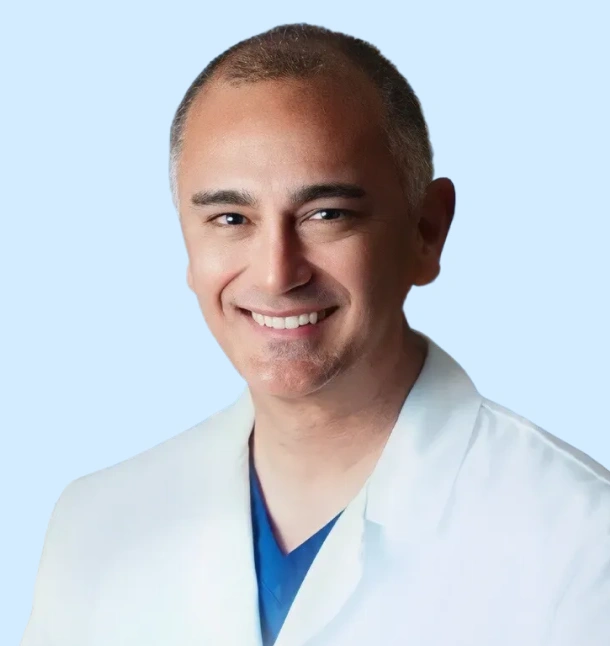 Male dentist in white coat and blue scrubs smiling at camera against light blue background.