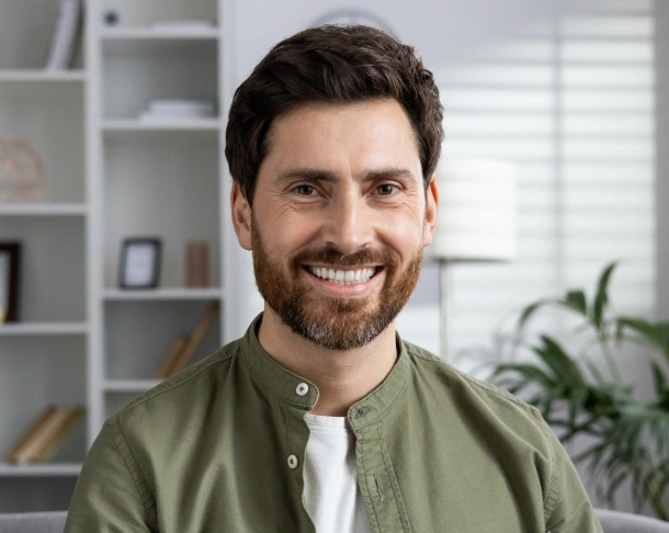 Smiling male dentist in olive green shirt at modern dental office with shelving and plant background.