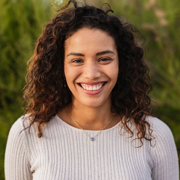Smiling woman with curly brown hair wearing light sweater outdoors with green background.