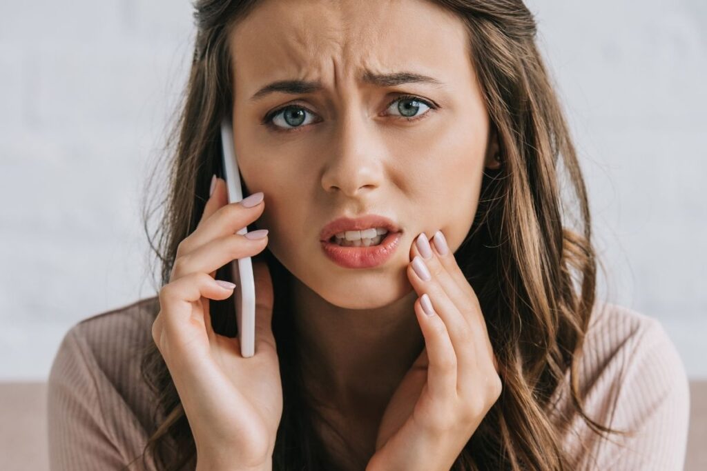 Concerned woman holding her jaw while talking on the phone, seeking dental help for sudden tooth pain or a dental emergency.