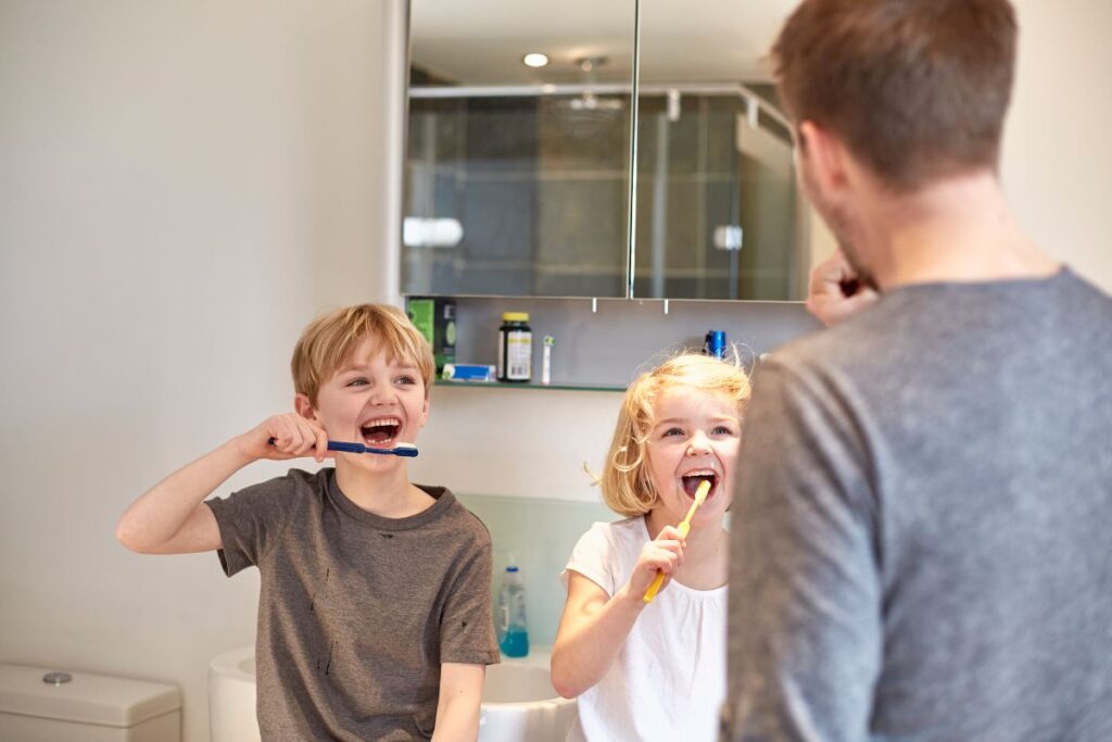 Two children happily brush their teeth together in front of a mirror while an adult guides them, promoting good oral hygiene habits.