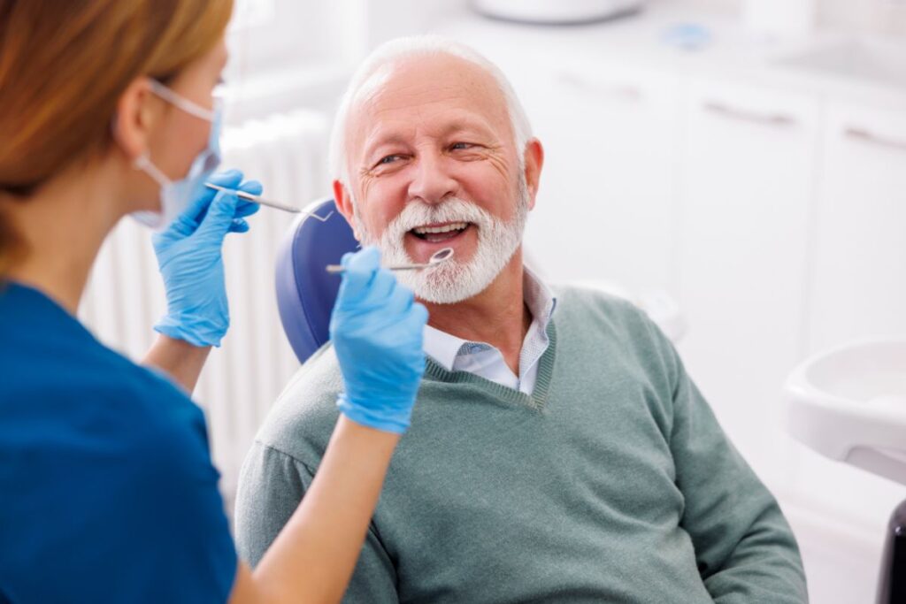 A dentist performs a routine dental check-up while speaking with an older patient during an implant care visit.