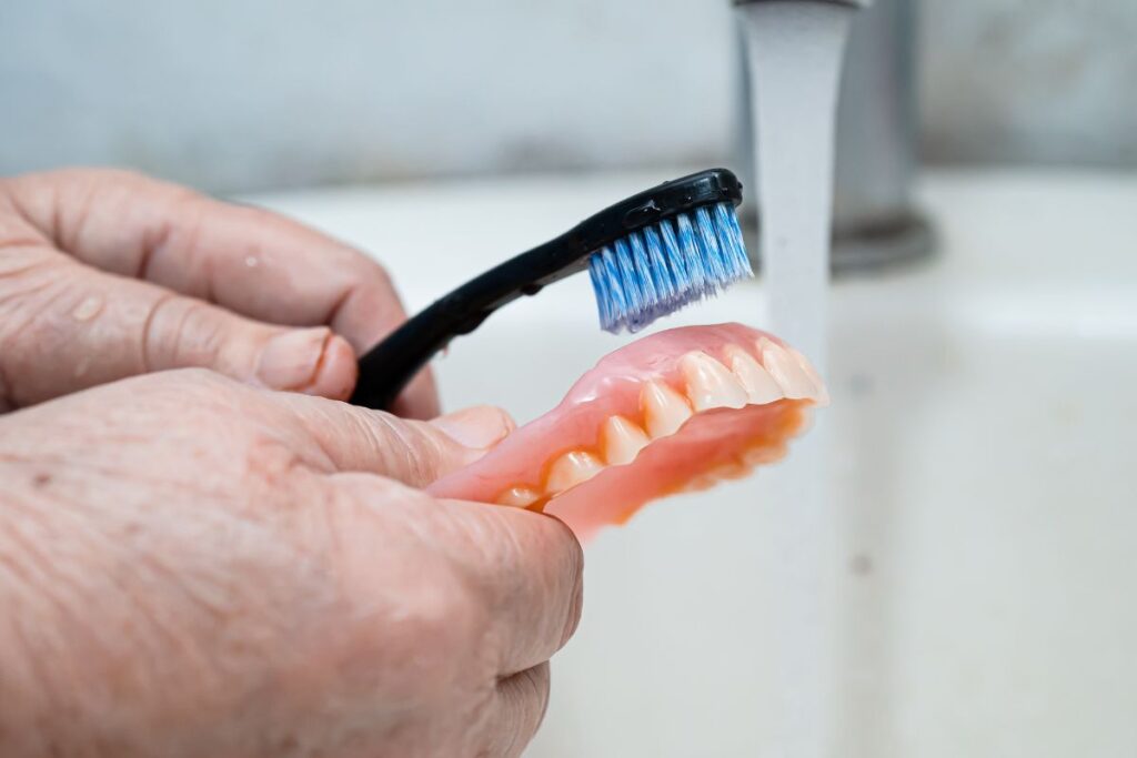 Person cleaning a removable denture with a toothbrush under running water, demonstrating proper denture care and maintenance.