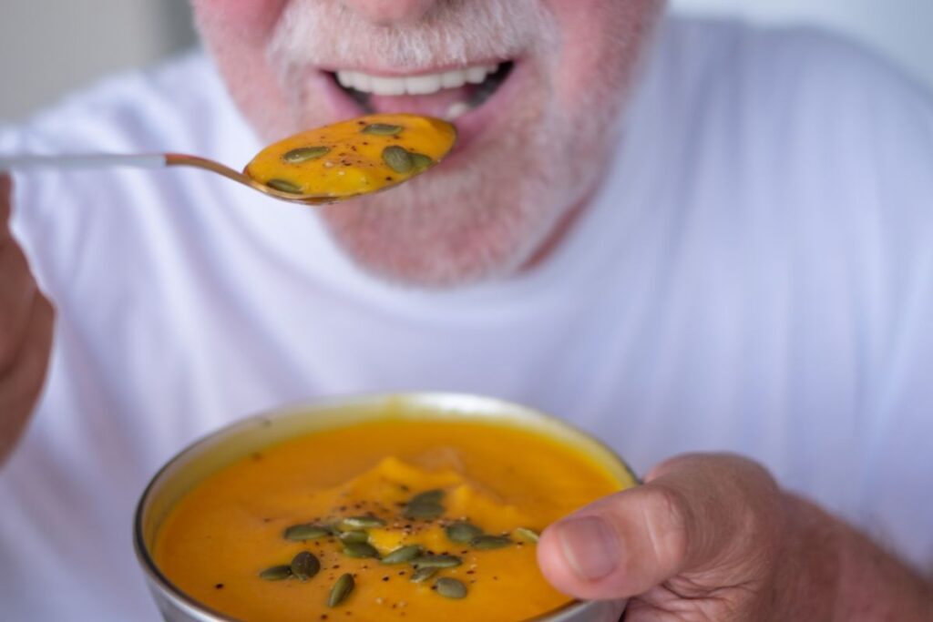 Older adult eating a bowl of soft soup, highlighting the adjustment period and recommended foods for new denture wearers.