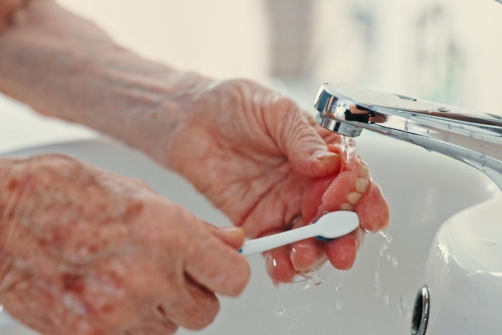 Senior adult brushing and cleaning removable dentures under running water